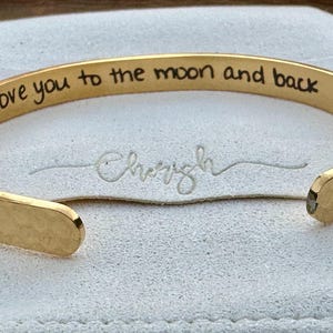 May include: Gold cuff bracelet with the inscription "I love you to the moon and back" engraved on the outside. The bracelet is on a white background.