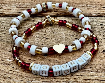 May include: Two beaded bracelets featuring white disc beads, metallic gold beads, and translucent red beads. One bracelet includes a gold heart charm, while the other features white square beads with gold lettering spelling GO HORNS.
