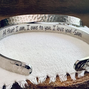 May include: A silver hammered bangle bracelet with an engraved message: "All that I am, I owe to you. I love you! - Sami"