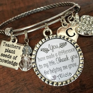 May include: Silver charm bracelet with a silver charm that says "Teachers plant seeds of knowledge". There is also a charm with a white background and black text that says "You have made a difference in my life. Thank you for helping me grow. Alicia". There is also a silver charm with a letter C and a silver charm with a rhinestone apple.
