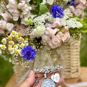 May include: A silver-toned brooch pin featuring a floral design, a round pendant with the text "Today a Bride, Tomorrow a Wife, Forever Our Little Girl, Mom & Dad", a heart-shaped charm with "Payton & Spencer 7/24/2025", and a small blue crystal bead.