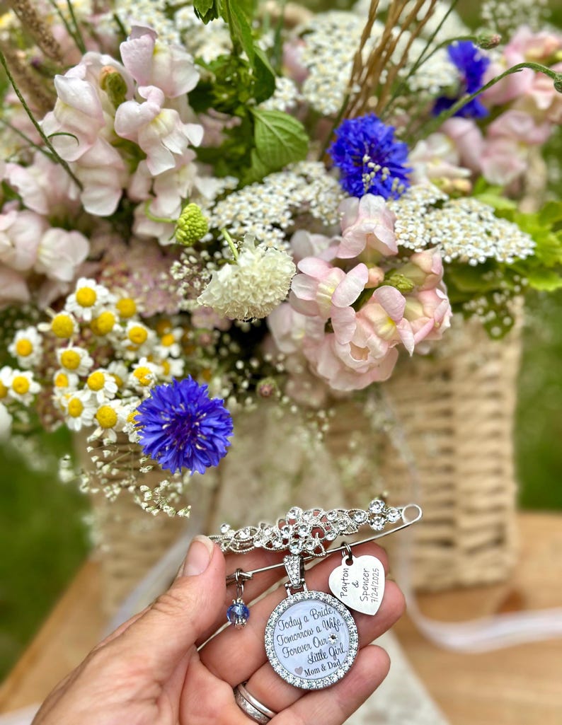 May include: A silver-toned brooch pin featuring a floral design, a round pendant with the text "Today a Bride, Tomorrow a Wife, Forever Our Little Girl, Mom & Dad", a heart-shaped charm with "Payton & Spencer 7/24/2025", and a small blue crystal bead.