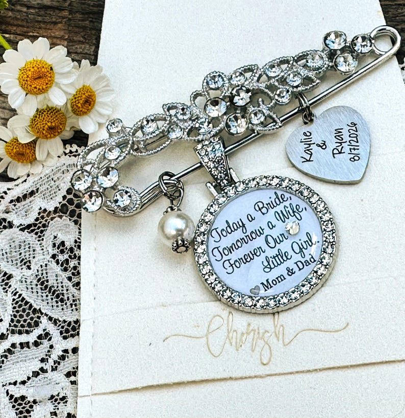 May include: A silver-toned brooch pin featuring clear rhinestones, a dangling white pearl bead, and two charms. One heart-shaped charm is engraved with "Kaylie & Ryan 8/7/2026". A round charm displays the text "Today a Bride, Tomorrow a Wife, Forever Our Little Girl. Mom & Dad".