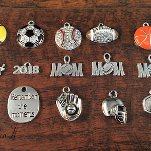 May include: A collection of 15 silver charms featuring various sports themes. The charms include a softball, soccer ball, baseball, football, basketball, a softball mom charm, a 2018 charm, three mom charms with different sports, a baseball glove, a football helmet, and a basketball hoop. The charms are perfect for adding a personal touch to jewelry or crafts.