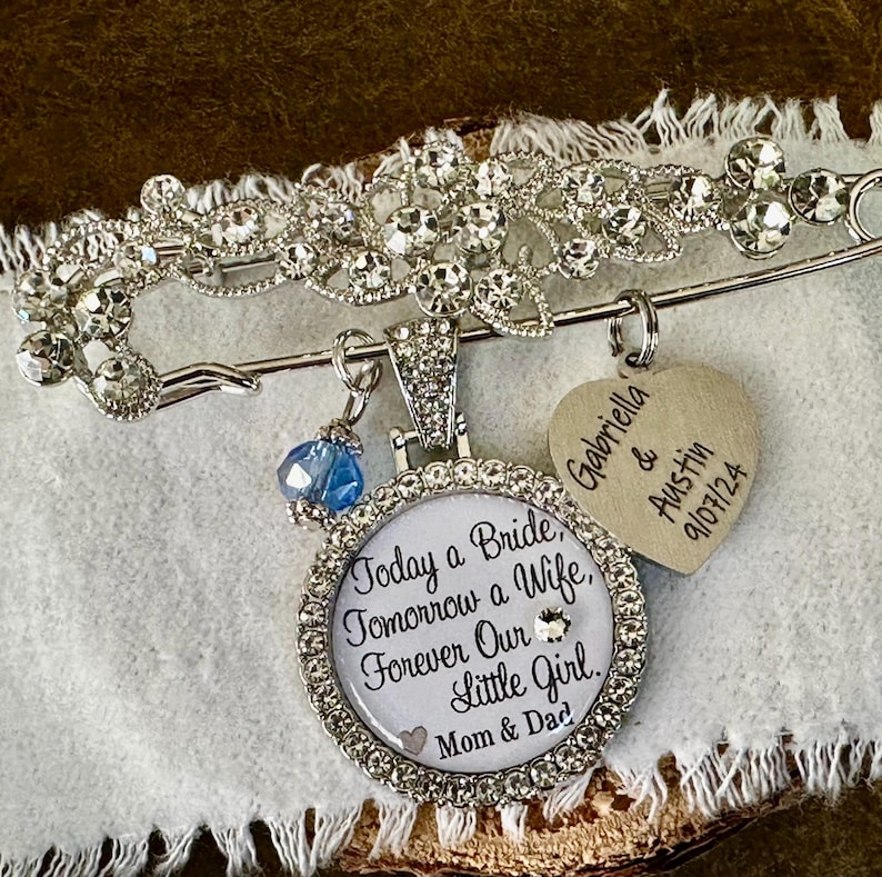 May include: A silver-toned brooch featuring a decorative filigree top set with clear rhinestones. A circular pendant hangs from the brooch, inscribed with the text Today a Bride, Tomorrow a Wife, Forever Our Little Girl, Mom & Dad. A heart-shaped charm is also attached, engraved with Gabriella & Austin 9/07/24, alongside a small blue bead.