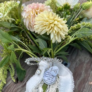 May include: A silver-toned brooch with clear rhinestones, a white pearl bead, and two charms attached to a white lace ribbon. One heart-shaped charm reads "Darby & Adel 10/06/26" and a round charm reads "Today a Tomorrow a Wife Forever Our Little Girl Mom & Dad".