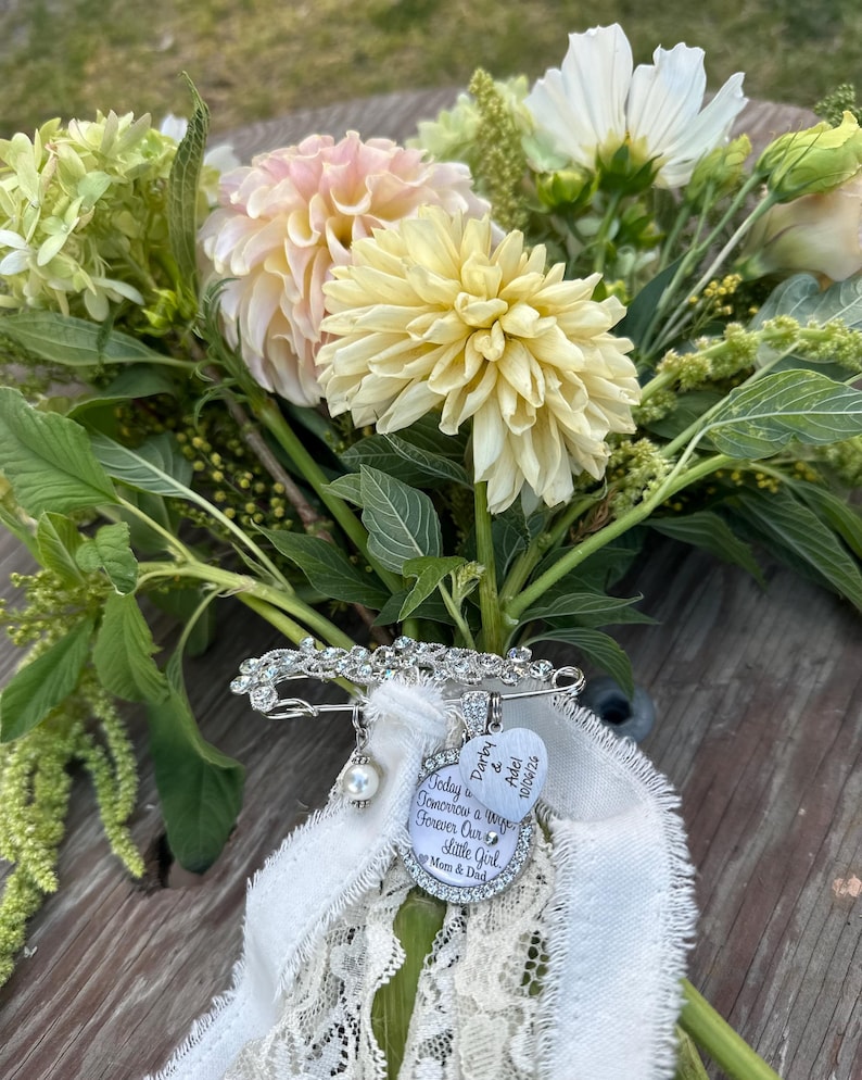 May include: A silver-toned brooch with clear rhinestones, a white pearl bead, and two charms attached to a white lace ribbon. One heart-shaped charm reads "Darby & Adel 10/06/26" and a round charm reads "Today a Tomorrow a Wife Forever Our Little Girl Mom & Dad".