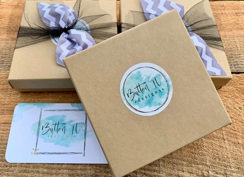 May include: Kraft-colored gift boxes with black mesh ribbon and chevron bows. Each box has a round sticker with the text "Button It designs." A business card with the same logo is also visible.