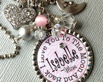 May include: Silver necklace with a pink and white charm that says "I love you to the moon and back - Mom" with the name "Isabelle" on it. The necklace also has a silver heart charm, a pink bead, and a silver crescent moon charm.