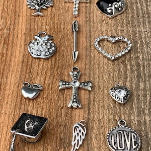 May include: Assortment of silver charm pendants, including a tree, cross, heart, apple, arrow, graduation cap, angel wing, and a circle with the word "LOVE".