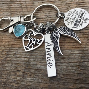 May include: Silver keychain with a blue charm, a heart charm with the word "Sweet 16" on it, a silver angel wing charm, and a silver tag with the name "Annie" engraved on it. The keychain also has a round charm with the text "Never Drive Faster Than Your Guardian Angel Can Fly".