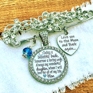 May include: A silver-toned brooch featuring clear rhinestones, a blue crystal bead, and two charms. One heart-shaped charm reads, Love you to the Moon and Back again. The circular charm reads, Today a beautiful bride, Tomorrow a loving wife, Always my wonderful daughter, whom I will love for all of my life, Mom.