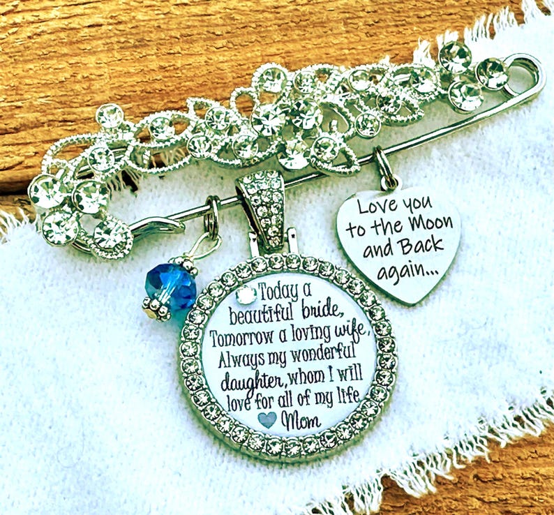 May include: A silver-toned brooch featuring clear rhinestones, a blue crystal bead, and two charms. One heart-shaped charm reads, Love you to the Moon and Back again. The circular charm reads, Today a beautiful bride, Tomorrow a loving wife, Always my wonderful daughter, whom I will love for all of my life, Mom.