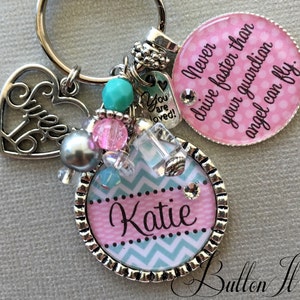 May include: A silver keychain with a pink and blue chevron patterned charm with the name "Katie" on it. The keychain also has a silver heart charm with the words "Sweet 16" on it, a silver charm with the words "You are loved!" on it, and a pink and white polka dot charm with the text "Never drive faster than your guardian angel can fly."