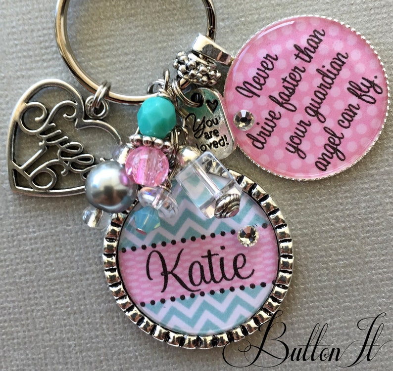 May include: A silver keychain with a pink and blue chevron patterned charm with the name "Katie" on it. The keychain also has a silver heart charm with the words "Sweet 16" on it, a silver charm with the words "You are loved!" on it, and a pink and white polka dot charm with the text "Never drive faster than your guardian angel can fly."
