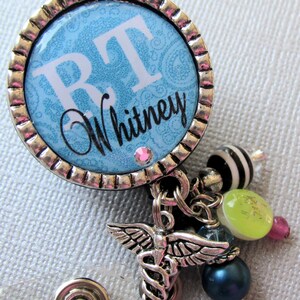 May include: A silver retractable badge holder with a blue and white patterned circle featuring the initials "RT" and the name "Whitney". The holder has a silver charm with a caduceus symbol and other charms including a green bead, a pink bead, and a black and white bead.