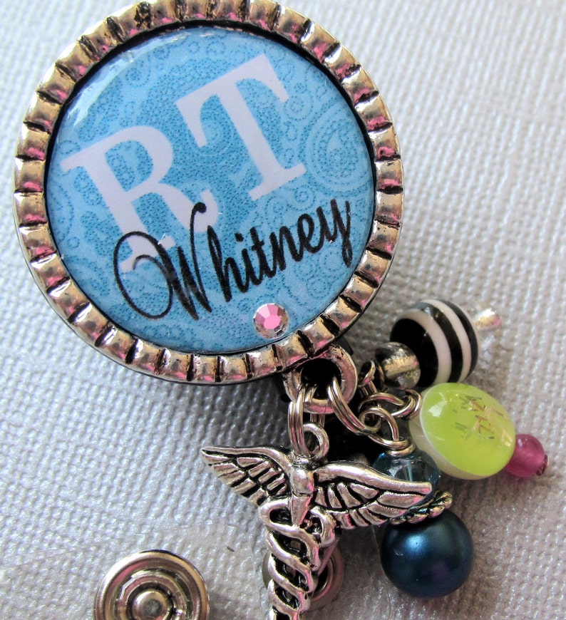 May include: A silver retractable badge holder with a blue and white patterned circle featuring the initials "RT" and the name "Whitney". The holder has a silver charm with a caduceus symbol and other charms including a green bead, a pink bead, and a black and white bead.