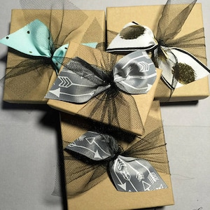 May include: Four small gift boxes wrapped in brown paper with black tulle and ribbon bows. The bows are tied with different patterned ribbon, including a light blue ribbon with black polka dots, a black and white ribbon with a white arrow pattern, and a black and white ribbon with a gray arrow pattern. The boxes are arranged in a cross shape.