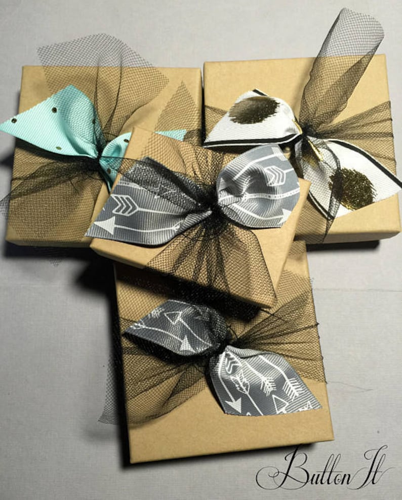 May include: Four small gift boxes wrapped in brown paper with black tulle and ribbon bows. The bows are tied with different patterned ribbon, including a light blue ribbon with black polka dots, a black and white ribbon with a white arrow pattern, and a black and white ribbon with a gray arrow pattern. The boxes are arranged in a cross shape.