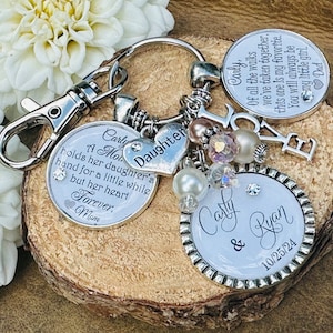 May include: A silver keychain with a charm that reads "A Mom's Daughter holds her daughter's hand for a little while but her heart forever, Mom".  Another charm reads "Daughter" and a third charm reads "Carly & Ryan 10/25/24".  The keychain also has a charm that reads "Love" and several silver and white beads.