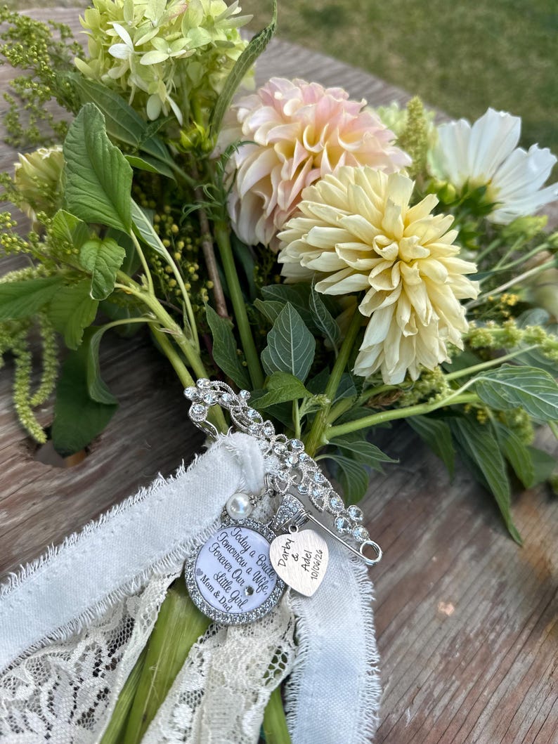 May include: A silver-toned brooch with clear crystals attached to a white lace ribbon, featuring a round charm inscribed with "Today a Bride, Tomorrow a Wife, Forever Our Little Girl, Mom & Dad" and a heart charm engraved with "Darby & Adel 10/06/26".