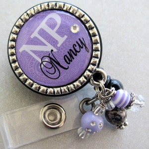 May include: Purple and silver badge holder with a clear retractable cord. The badge has a purple background with a white "NP" and the name "Nancy" in black script. The badge holder has a silver charm with purple and white striped beads.