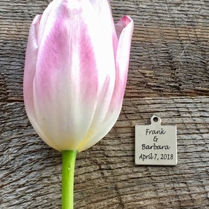 May include: A pink tulip with a green stem sits on a wooden surface next to a silver square tag with the text "Frank & Barbara April 7, 2018" engraved on it.