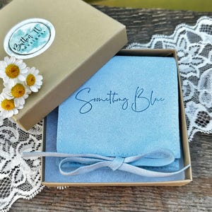 May include: A light blue suede jewelry pouch with the text Something Blue printed in dark blue script, tied with a matching ribbon, nestled inside a square cardboard gift box featuring a Button IV designs sticker and white daisy flowers.