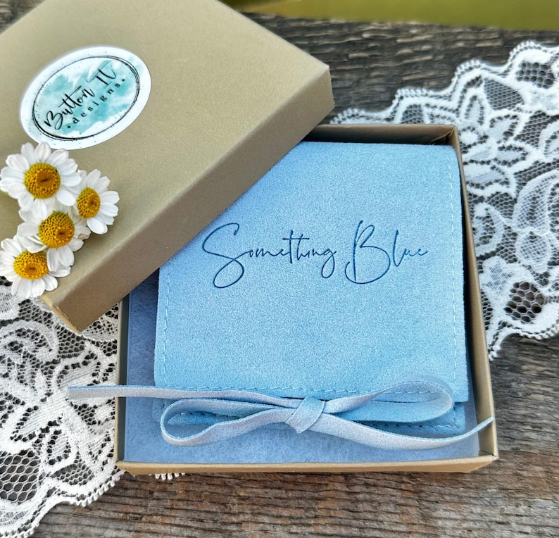 May include: A light blue suede jewelry pouch with the text Something Blue printed in dark blue script, tied with a matching ribbon, nestled inside a square cardboard gift box featuring a Button IV designs sticker and white daisy flowers.