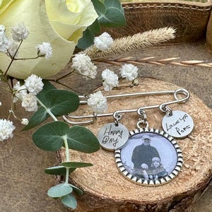 May include: A silver brooch with a safety pin design, featuring three circular charms. One charm has a photo, while the others read "Happy Day" and "Love Ya Pops". The brooch is displayed on a wooden surface, surrounded by flowers.