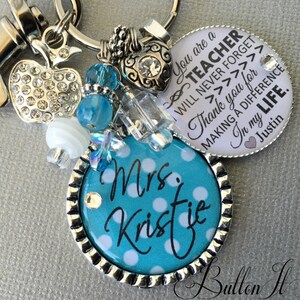May include: A teal blue and white polka dot keychain with the text "Mrs. Kristie" on it. The keychain also has a silver charm with a heart and rhinestones, a silver apple charm with rhinestones, and a silver charm with the text "You are a Teacher I will never forget Thank you for making a difference in my life. Justin".