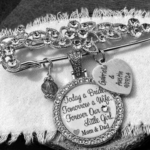 May include: A silver-toned brooch pin featuring a decorative floral top encrusted with clear crystals. It includes a round pendant with the text Today a Bride, Tomorrow a Wife, Forever Our Little Girl, Mom & Dad, a heart charm with Gabriella & Austin 9/07/24, and a small crystal bead.