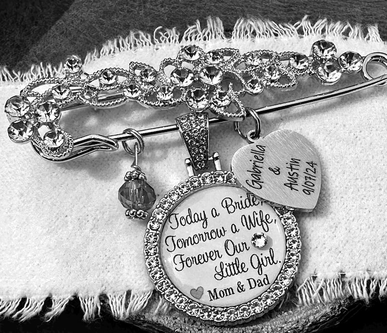 May include: A silver-toned brooch pin featuring a decorative floral top encrusted with clear crystals. It includes a round pendant with the text Today a Bride, Tomorrow a Wife, Forever Our Little Girl, Mom & Dad, a heart charm with Gabriella & Austin 9/07/24, and a small crystal bead.