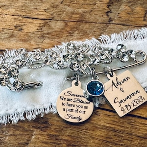 May include: A silver brooch with rhinestones and a charm that says "Savanna We are Blessed to have you as a part of our Family". There is also a charm with a blue rhinestone and a square charm that says "Adrian & Savanna 8-15-2024".