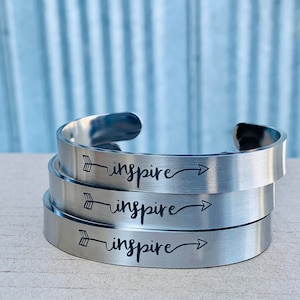 May include: Three silver-colored cuff bracelets stacked on top of each other. Each bracelet has the word "inspire" in a cursive font, with an arrow design. The bracelets have a brushed metal finish.