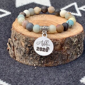 May include: A graduation bracelet with a silver charm that says "Ashlei 2020". The bracelet is made of natural stones in shades of brown, beige, and blue.