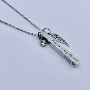 May include: Silver necklace with a bar pendant engraved with "Forever in my Heart Dad" and a small crystal charm. The bar pendant is attached to a silver chain with a small angel wing charm.