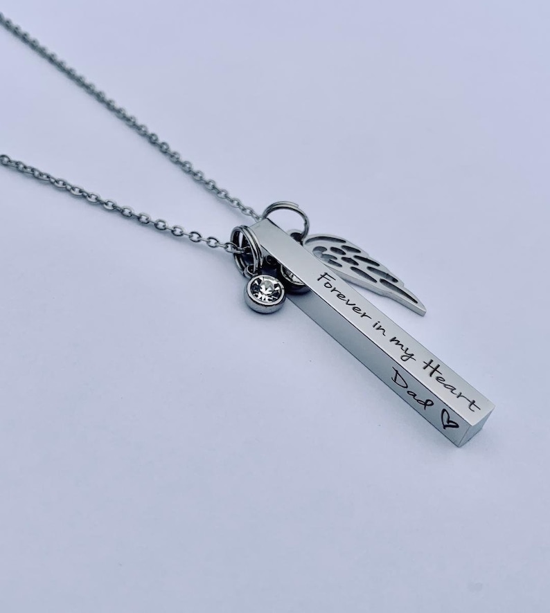 May include: Silver necklace with a bar pendant engraved with "Forever in my Heart Dad" and a small crystal charm. The bar pendant is attached to a silver chain with a small angel wing charm.