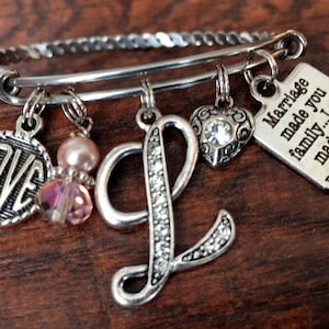 May include: A silver charm bracelet with a heart charm, a letter L charm, a charm that says "LOVE", a pink pearl charm, a pink crystal charm, and a square charm that says "Marriage made you family...I love you made you my daughter".