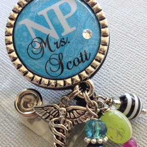 May include: A blue and silver badge holder with a white background and the text "NP Mrs. Scott". The badge holder has a silver butterfly charm with a snake wrapped around it, and a variety of colourful beads.