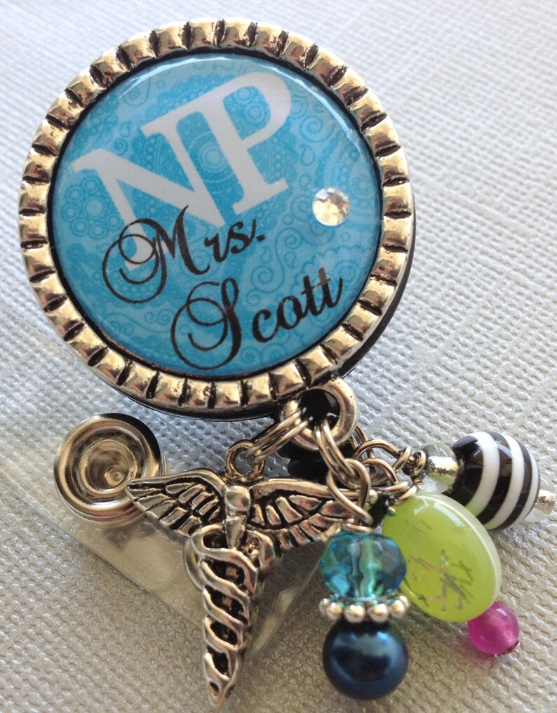 May include: A blue and silver badge holder with a white background and the text "NP Mrs. Scott". The badge holder has a silver butterfly charm with a snake wrapped around it, and a variety of colourful beads.
