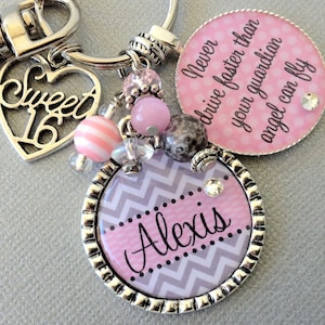 May include: A silver keychain with a pink and gray chevron patterned charm that says "Alexis" and a pink charm with the text "Never drive faster than your guardian angel can fly". The keychain also has a silver heart charm with the text "Sweet 16" and pink and white beads.