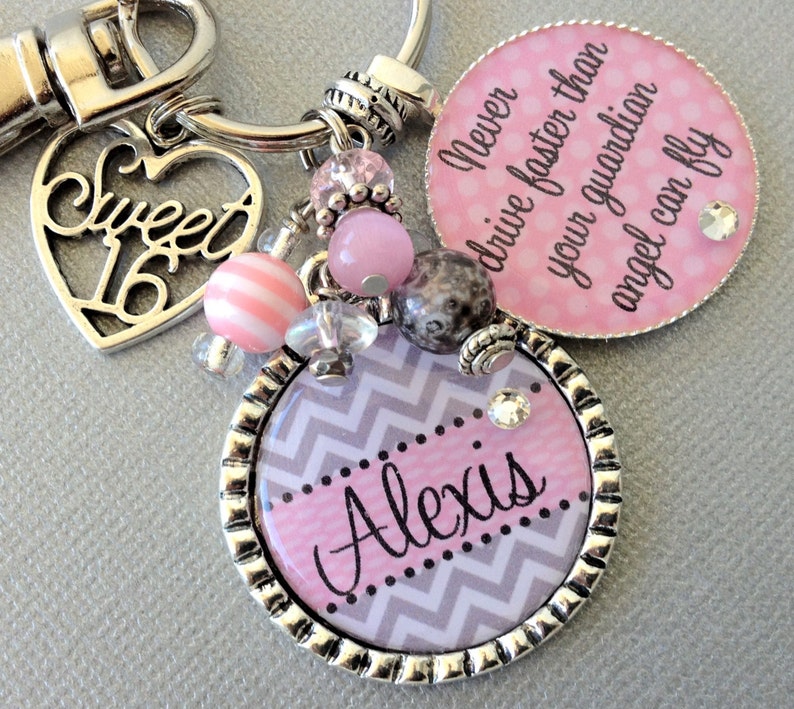 May include: A silver keychain with a pink and gray chevron patterned charm that says "Alexis" and a pink charm with the text "Never drive faster than your guardian angel can fly". The keychain also has a silver heart charm with the text "Sweet 16" and pink and white beads.