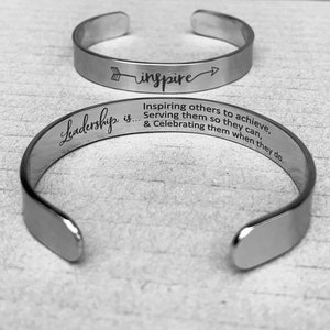 Puede incluir: Pulsera de plata con las palabras "Leadership is... Inspiring others to achieve, Serving them so they can, & Celebrating them when they do." y una segunda pulsera con una flecha y la palabra "inspire" en ella.