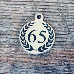 May include: A silver round charm with a black laurel wreath design and the number 65 in the center.