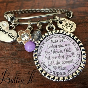 May include: A silver charm bracelet with a purple and white charm that reads "Here is something old to carry on your wedding day. Today you are the Flower Girl, but one day you'll hold the bouquet. Mom". The bracelet also has a silver heart charm that reads "Flower Girl", a silver flower charm with a purple center, and a silver charm that reads "You are loved!"