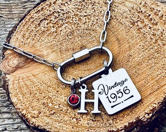 May include: A silver-toned necklace featuring a carabiner-style clasp as a pendant. Attached to the clasp are three charms: a small circular red gemstone, a silver letter H, and a square silver tag engraved with the text Vintage 1956.