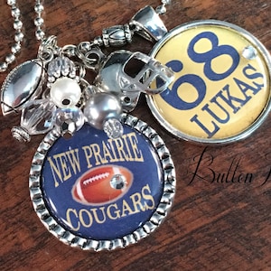 May include: A silver chain necklace with a variety of charms, including a football, a helmet, a pearl, and two round charms. One charm is yellow with the number 68 and the name "Lukas" in blue lettering. The other charm is blue with the words "New Prairie Cougars" and a football graphic.