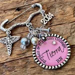 May include: A silver-toned brooch with a pin closure, featuring a medical symbol charm, beads, and a charm that reads "2024." A pink circular pendant displays the name "Tierra" and "RN" with "Bryant & Stratton College" around the edge.