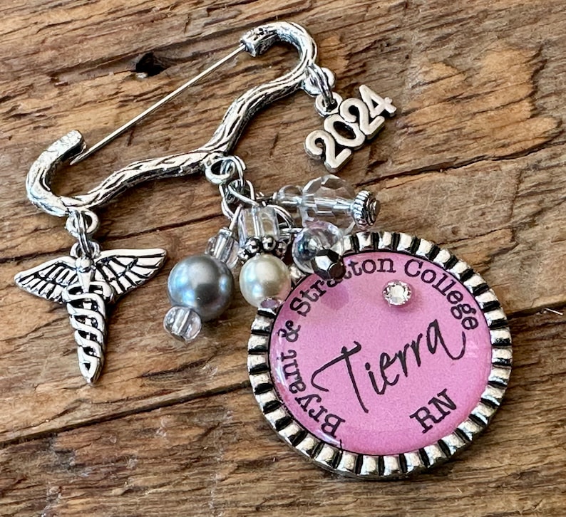 May include: A silver-toned brooch with a pin closure, featuring a medical symbol charm, beads, and a charm that reads "2024." A pink circular pendant displays the name "Tierra" and "RN" with "Bryant & Stratton College" around the edge.